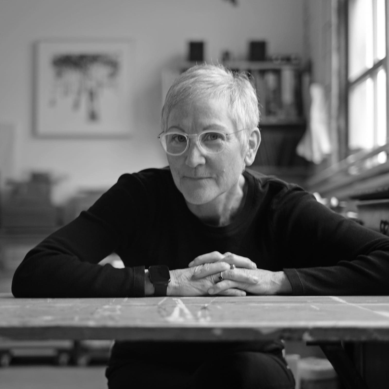 Black and white portrait of artist Maria Josenhans seated at a work table in her studio, hands clasped, looking directly at the camera.