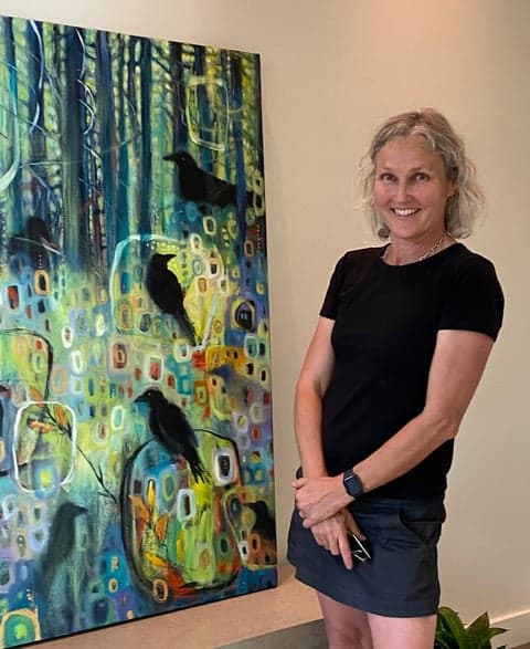 Woman standing indoors beside a large, colorful painting featuring crows in a forest-like scene, smiling toward the camera.