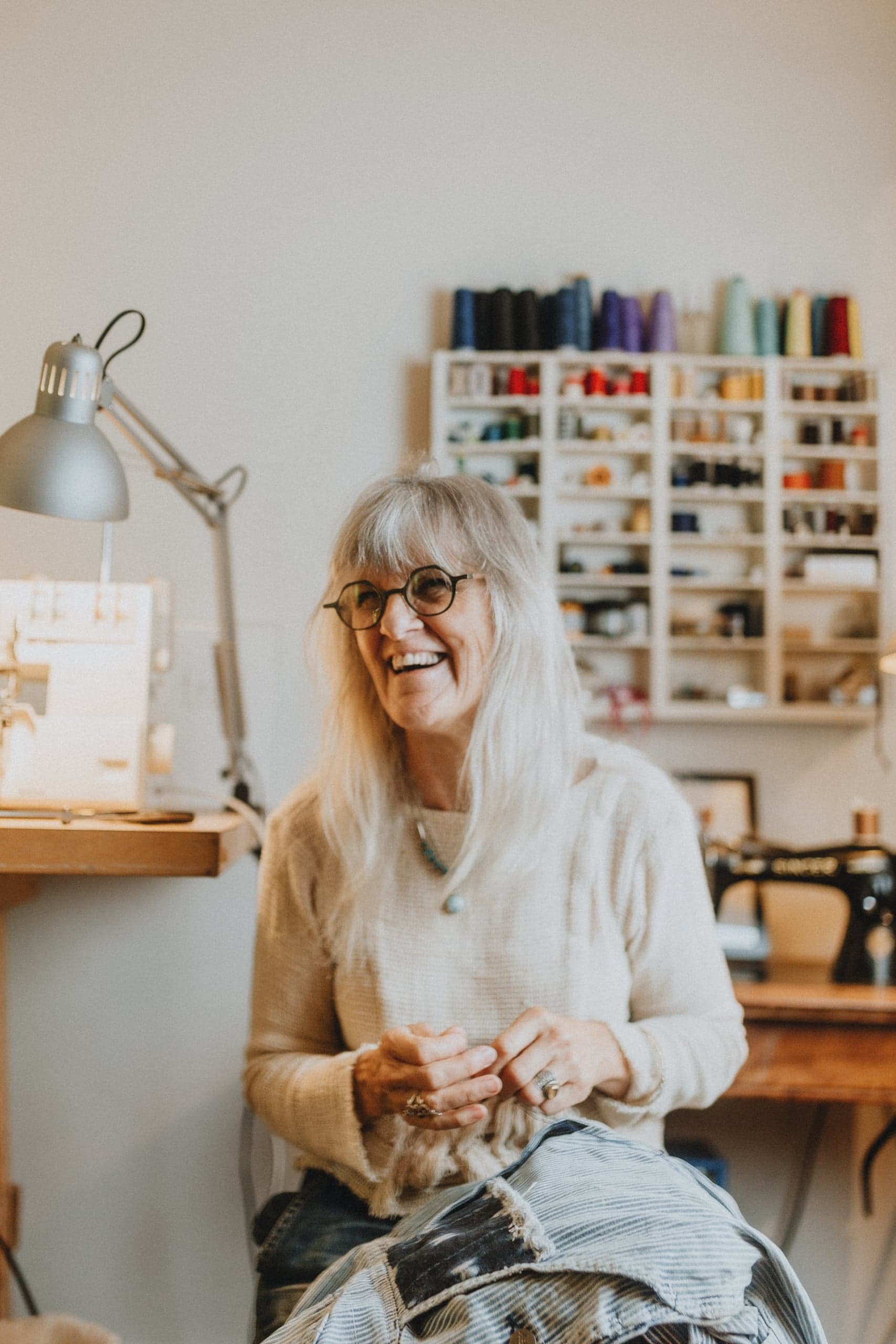 Woman with long gray hair and glasses smiling while sewing fabric in a bright studio with shelves of thread behind her.