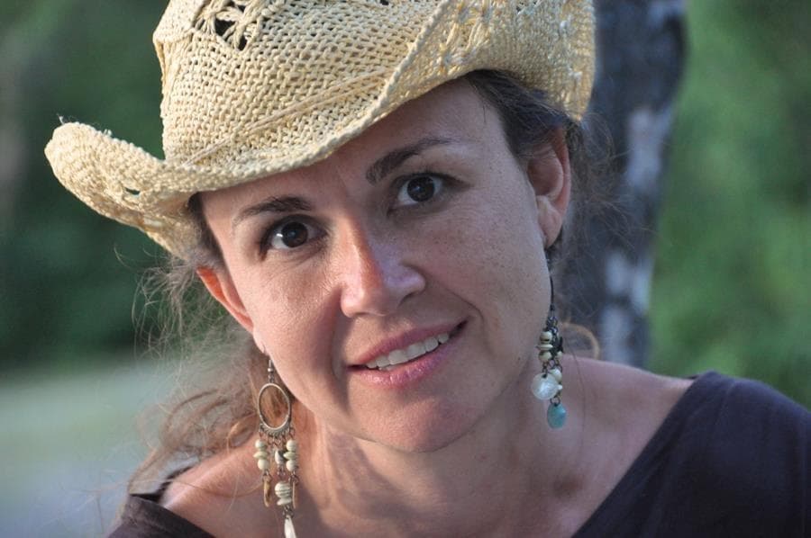 Woman wearing a woven straw hat and dangling earrings, smiling gently while looking toward the camera outdoors.