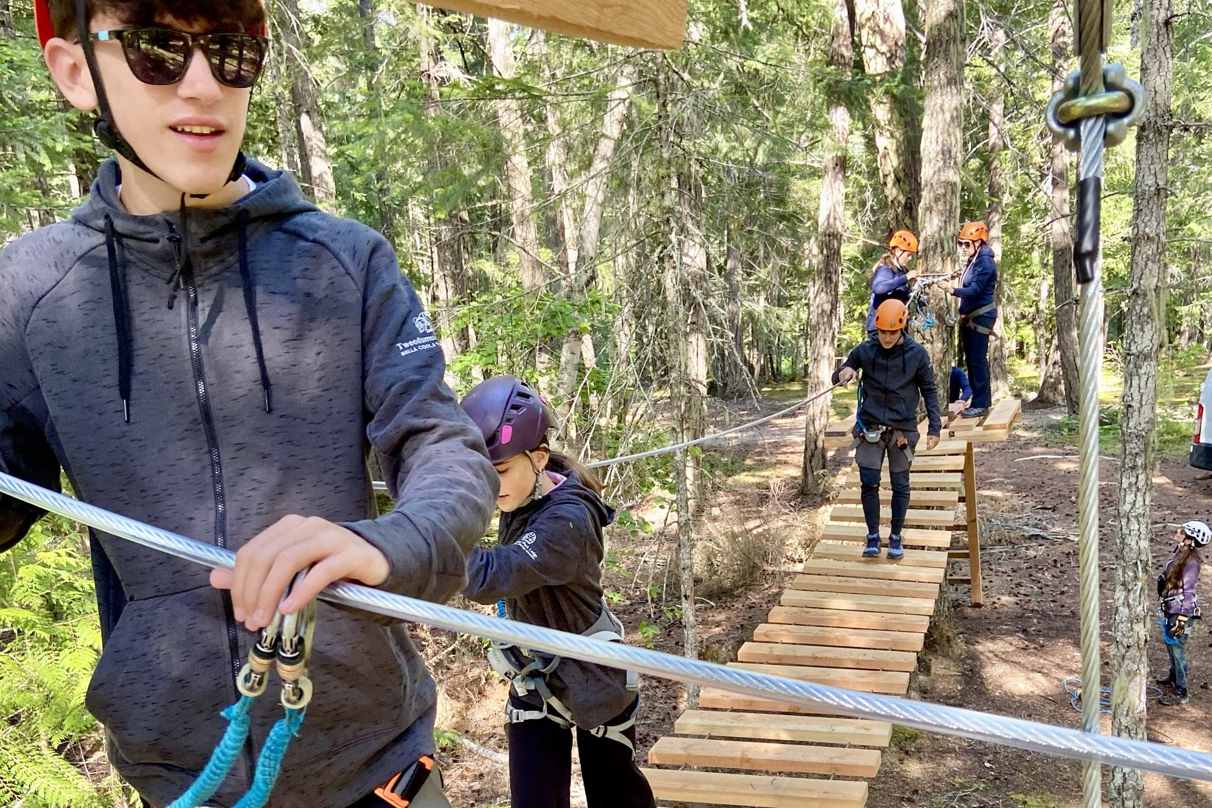 Participants wearing helmets and safety harnesses navigate a suspended wooden bridge on an aerial ropes course in a forest, while others wait and receive instructions on a platform in the background.