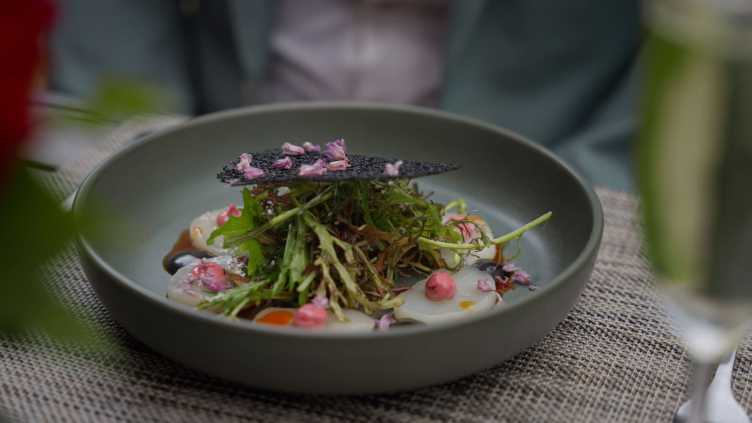 Plated gourmet salad with delicate greens, thinly sliced white seafood, edible flowers, and a crisp black garnish in a shallow bowl.